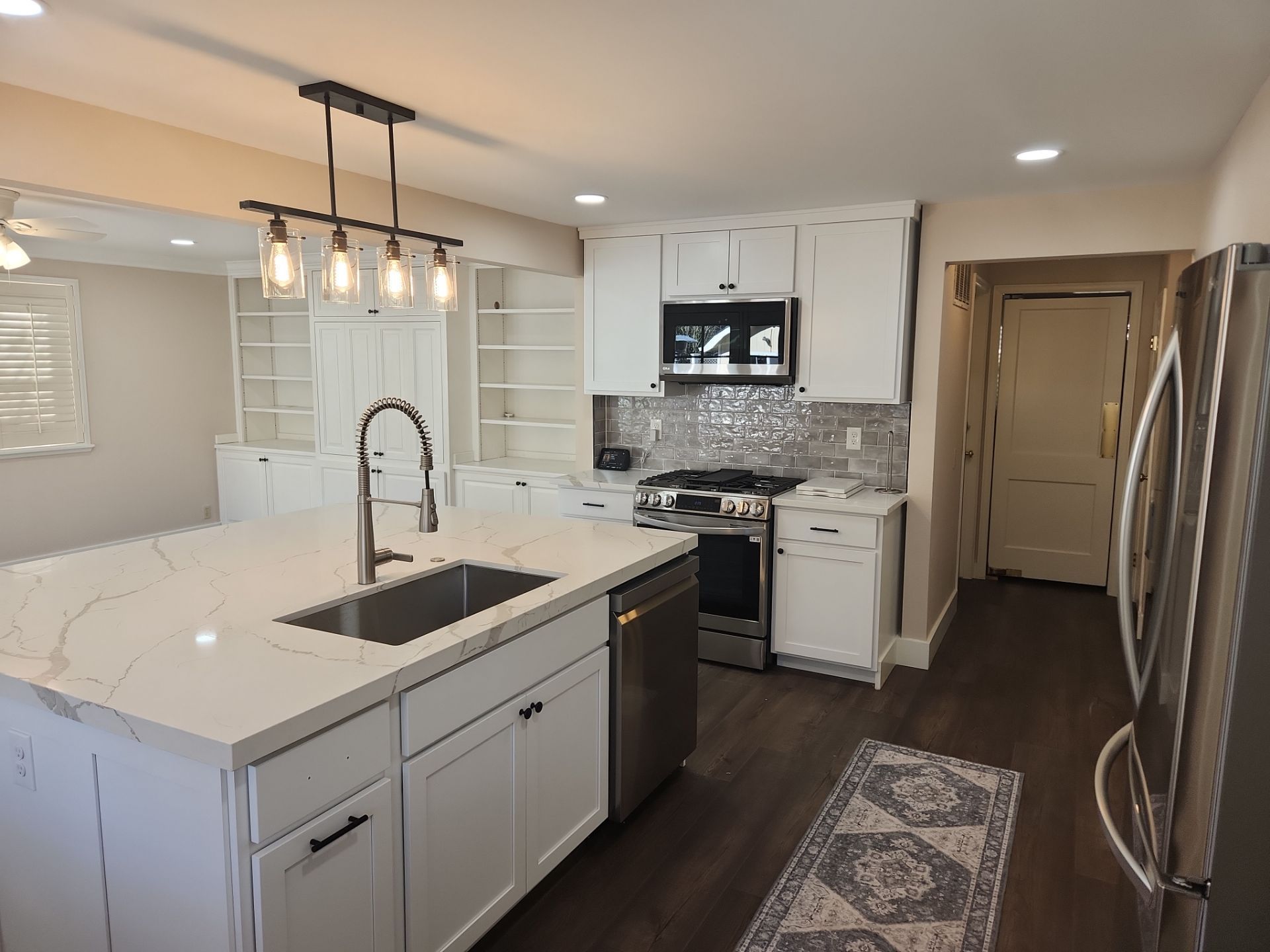 Modern kitchen with white cabinets, marble island, pendant lights, dark wood floor, and stainless steel fridge.