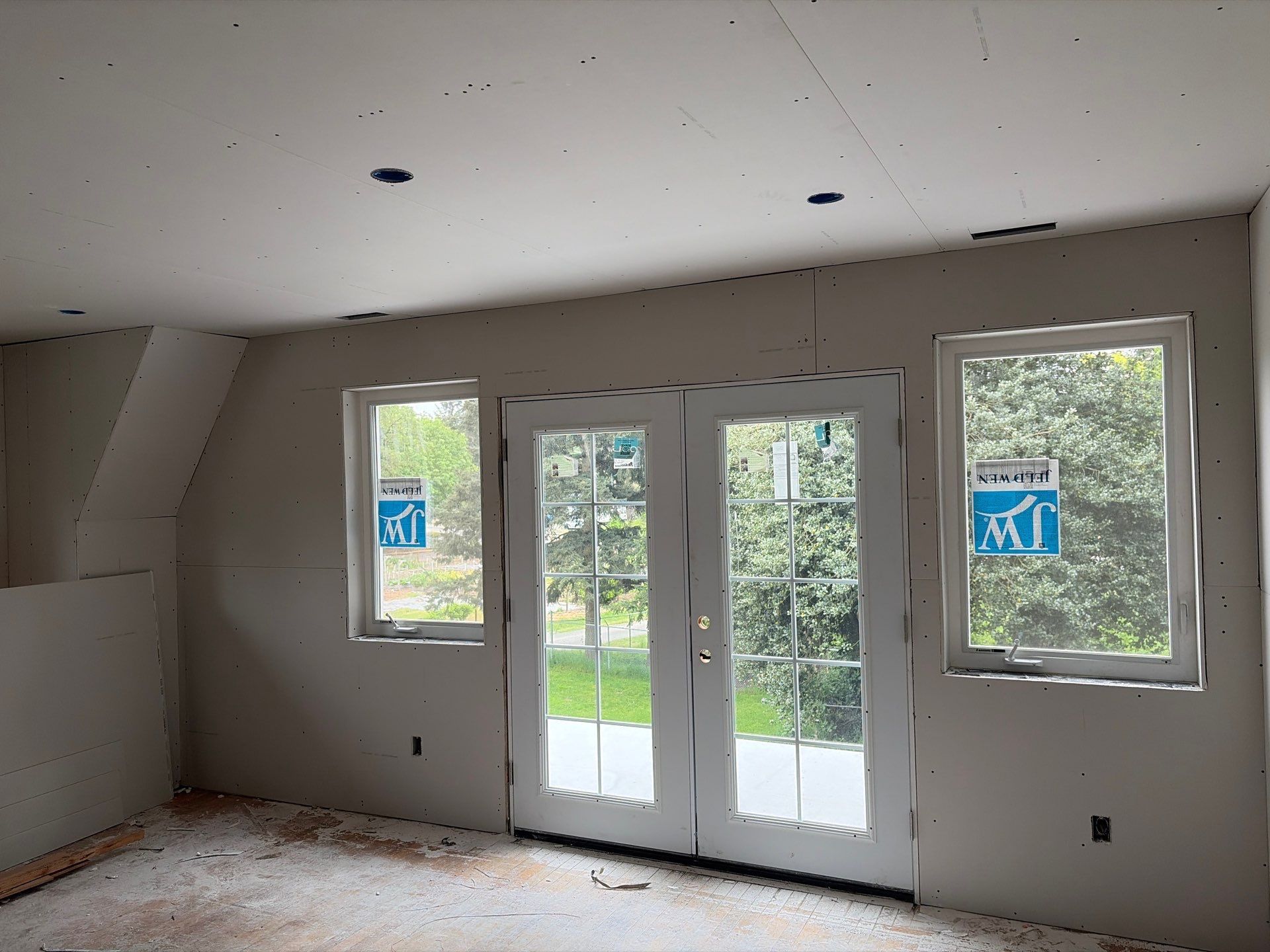 Empty room with white walls, windows, and French doors opening to a green yard.