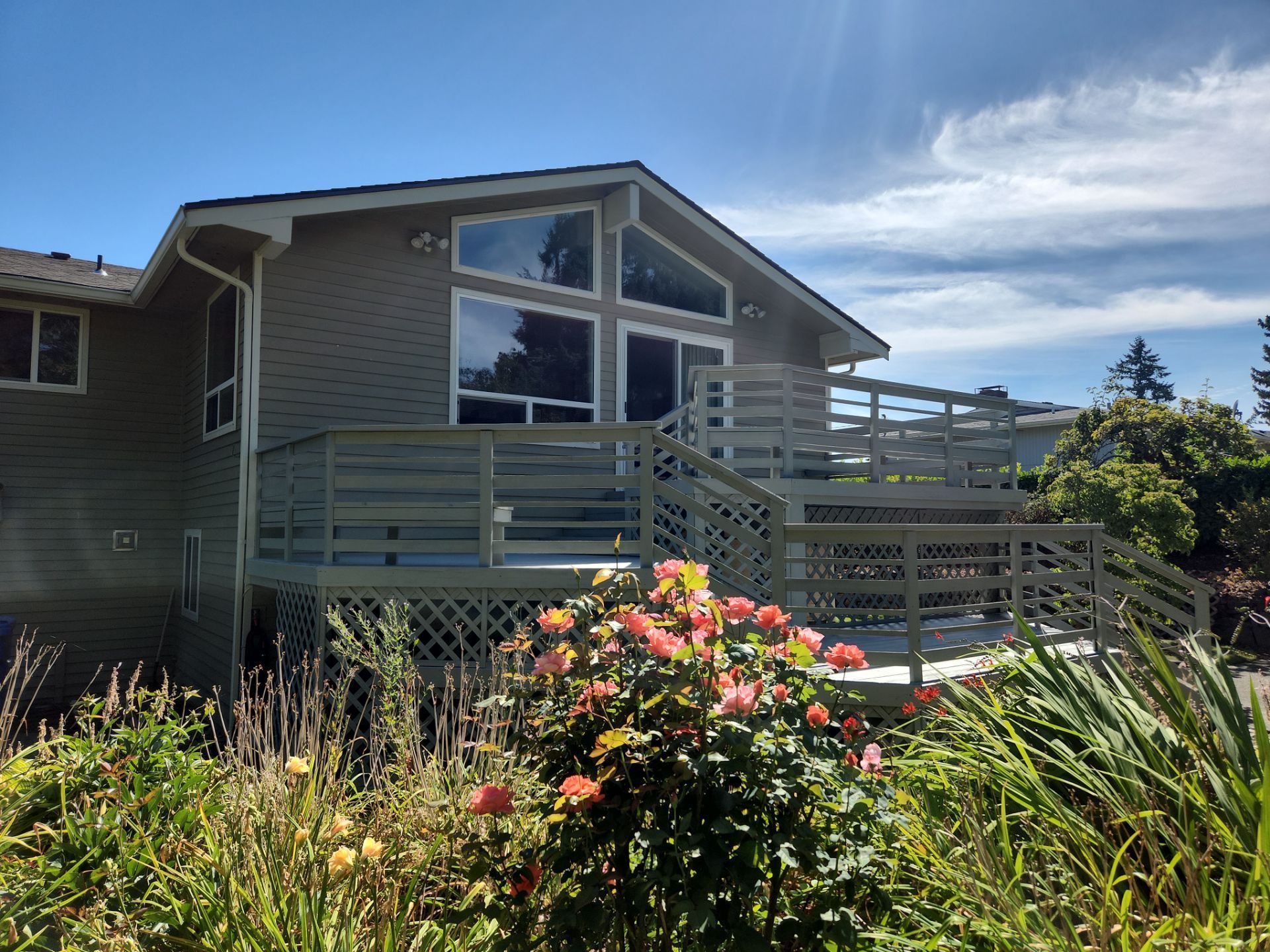 House with large windows and wooden deck, surrounded by tall grass and flowering shrubs under a cloudy sky