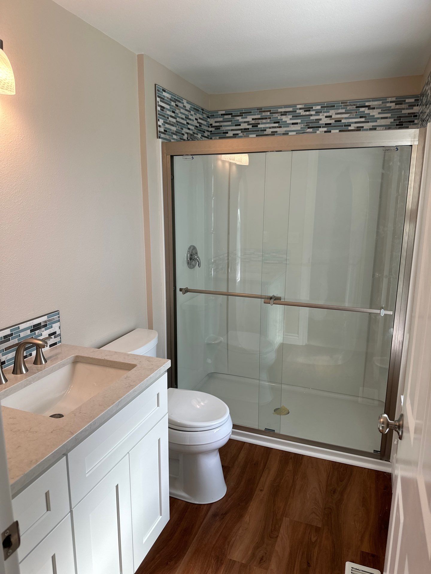 Small bathroom with white vanity, toilet, and glass shower with mosaic tile border and wood-look floor