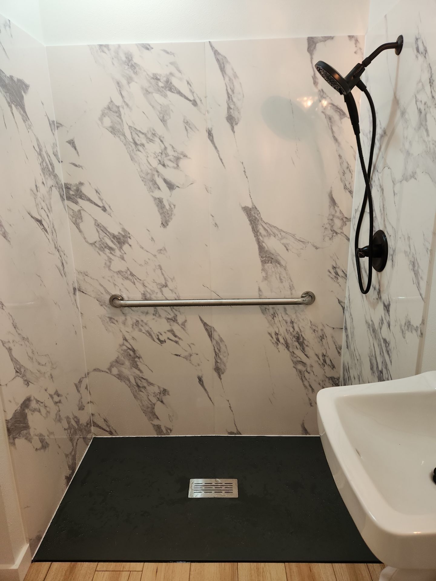 Walk-in shower with marble-look walls, black shower mat, grab bar, and handheld showerhead