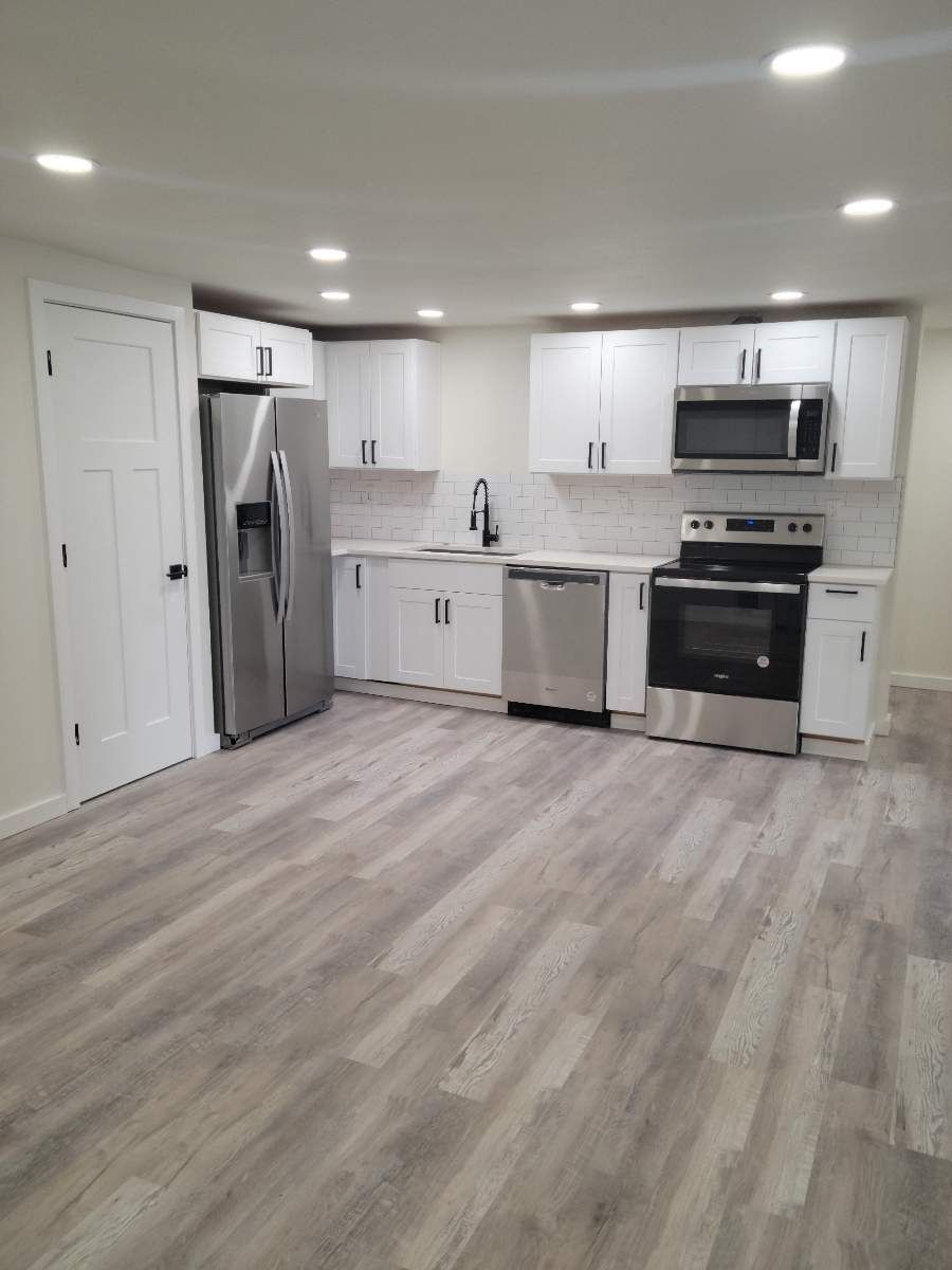 Modern white kitchen with stainless steel appliances and light wood flooring, viewed from an empty open room