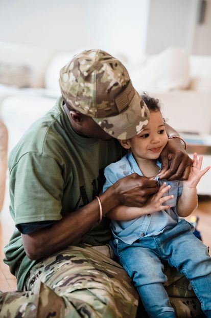 A soldier in camouflage gently holds a smiling child indoors on a couch.