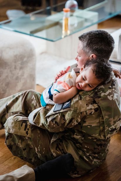Soldier in camouflage sitting indoors while holding a sleeping child on his lap
