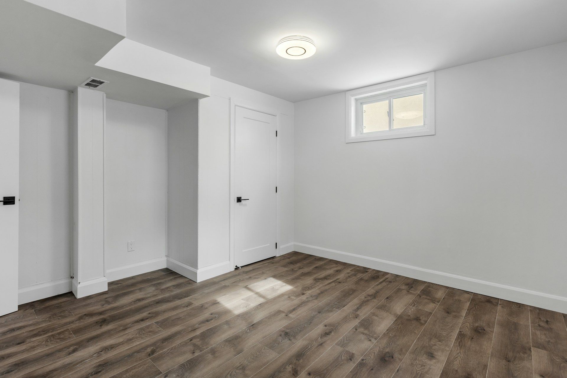 Empty white bedroom with wood flooring and a small window near the ceiling.