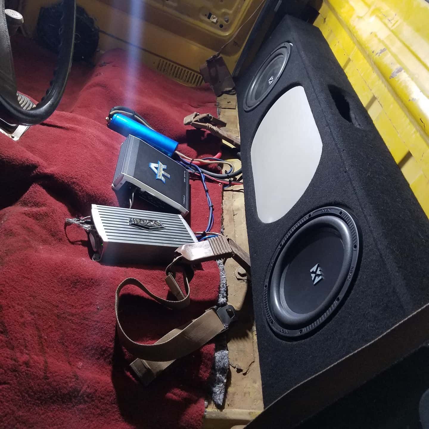 Interior of a yellow vehicle with red carpet. A subwoofer box, amplifiers, and other audio equipment are visible.