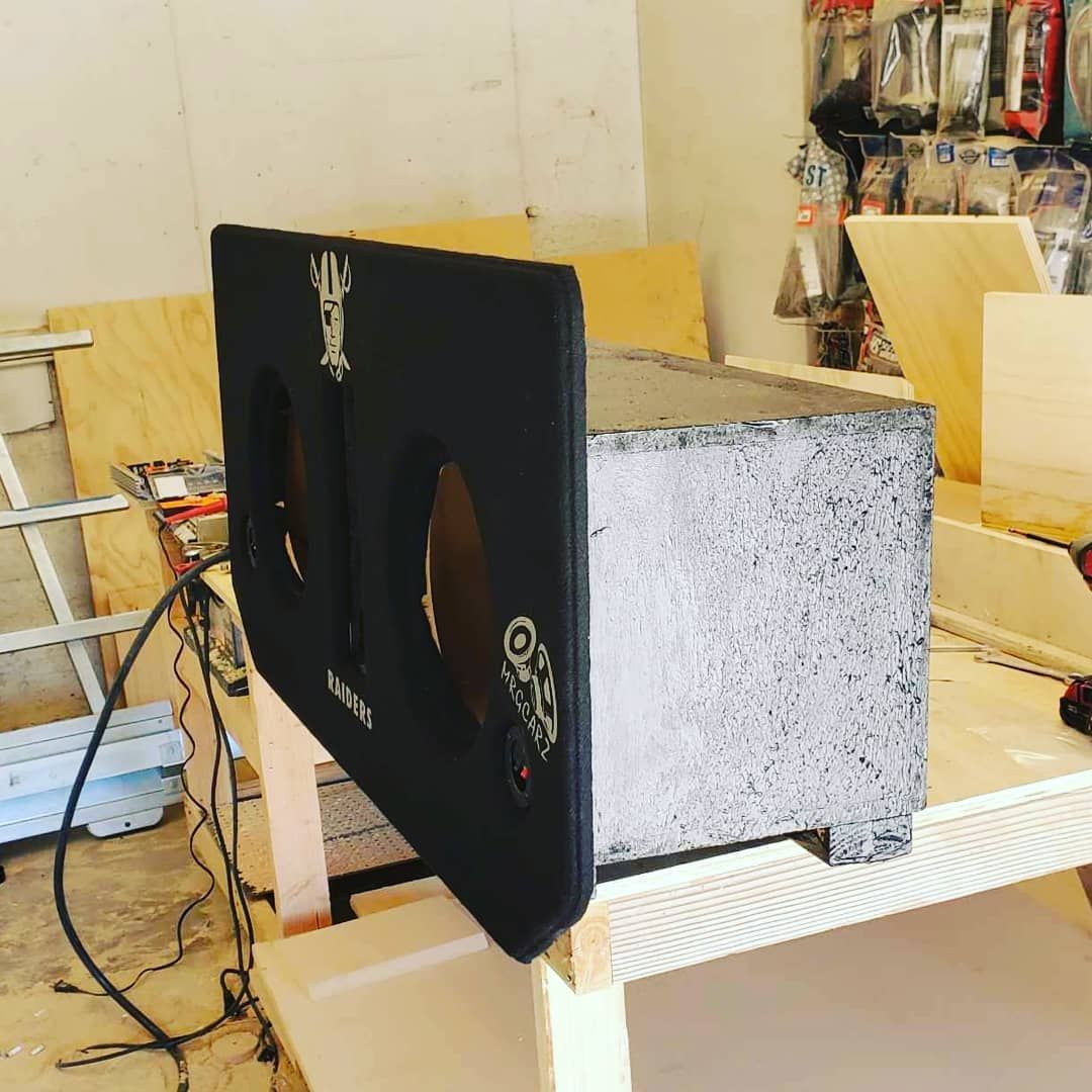 Black subwoofer box with two circular speaker holes, resting on a wooden table in a workshop setting.