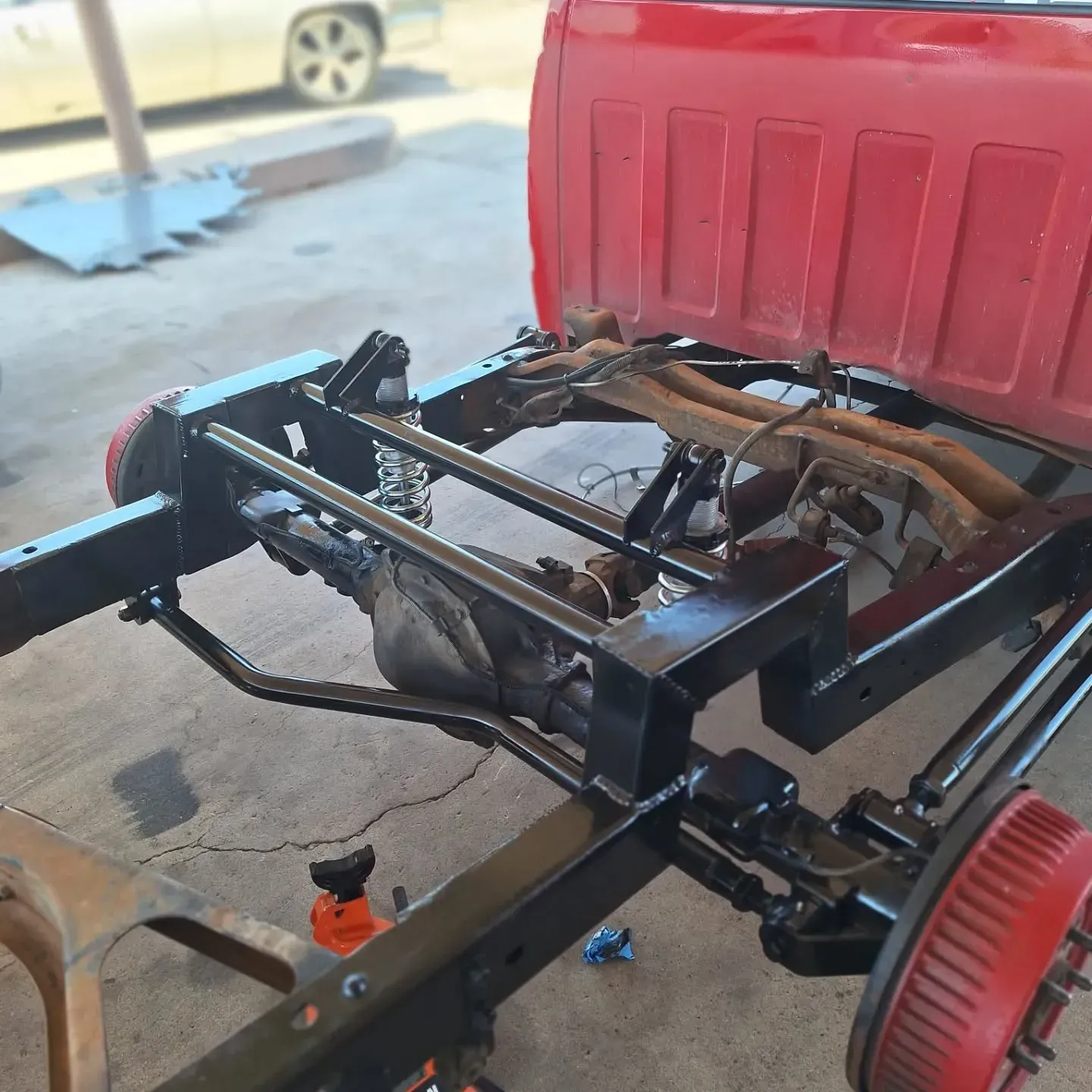 Red pickup truck chassis, painted black. The frame is supported on jack stands, with red brake drums visible.
