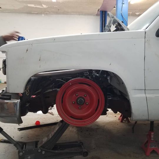 A white pickup truck with a red wheel, propped up on a jack in a garage. Someone is working on the truck.