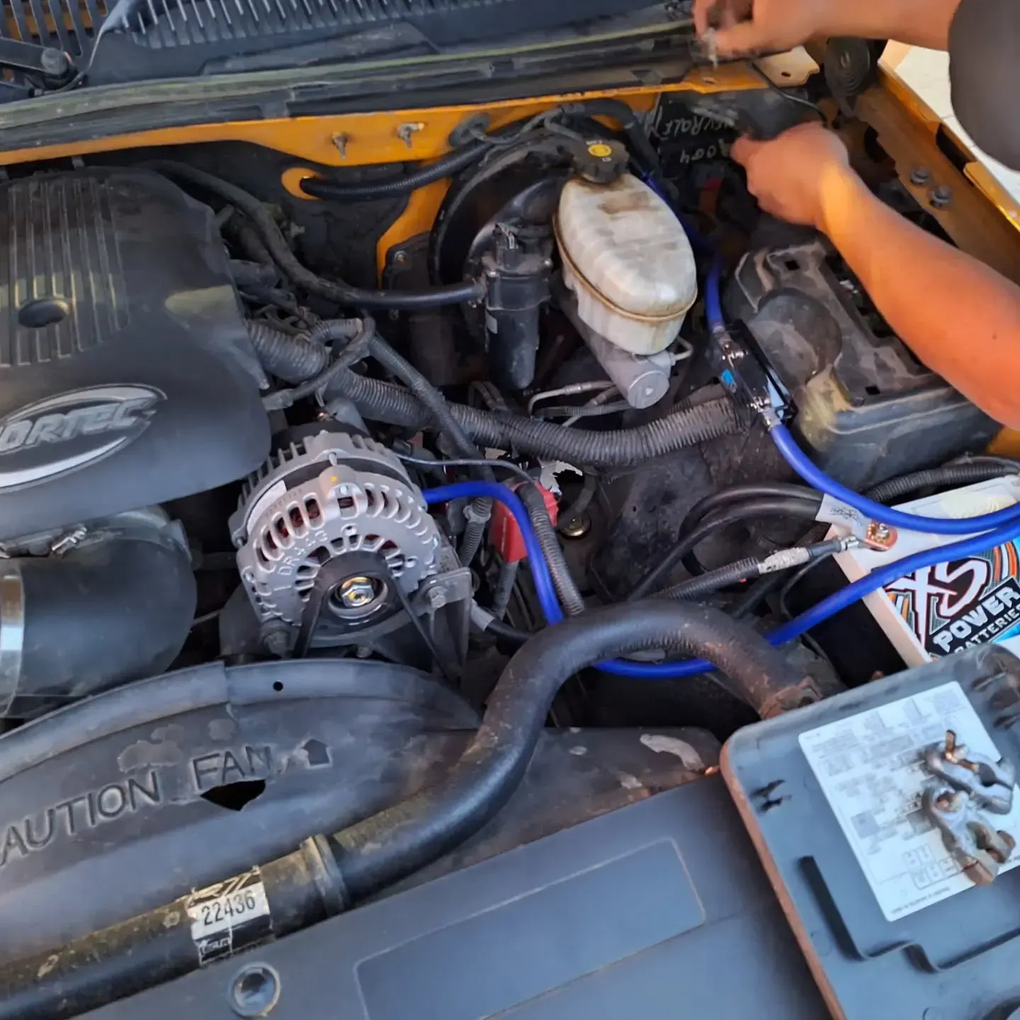 Person working on a yellow car engine, with a battery and hoses visible. The setting appears to be outdoors.