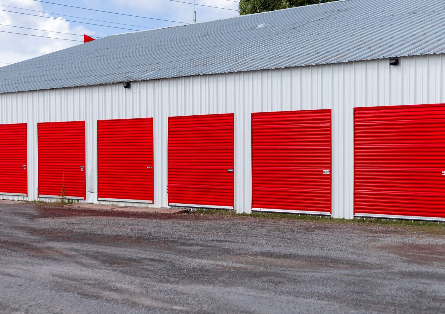 Red storage unit doors on a white building with a gray roof and a gravel driveway.