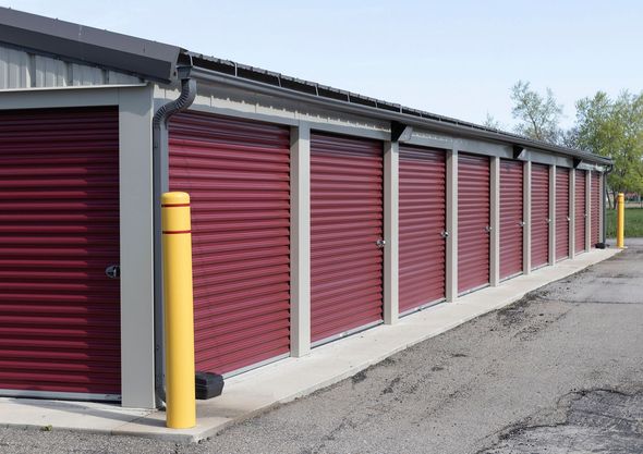 Storage units with burgundy roll-up doors, concrete base, gray frames, yellow bollards, and a light-colored asphalt surface. Storage units with burgundy roll-up doors, concrete base, gray frames, yellow bollards, and a light-colored asphalt surface.