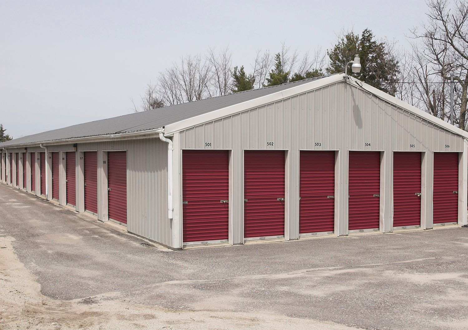 Storage units with red doors, gray metal building. Gravel driveway, outdoor setting.
