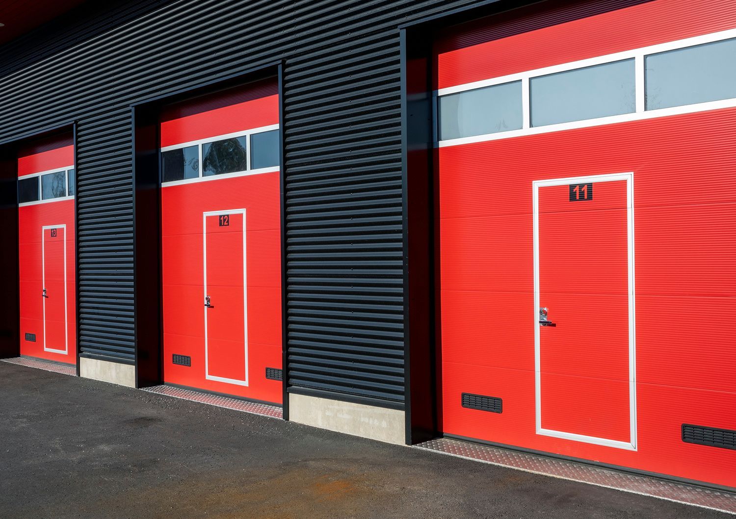 Three red garage doors with windows and a small pedestrian door built-in, set against a dark, corrugated wall.