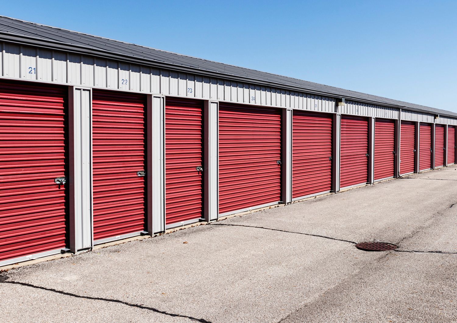 Red storage unit doors in a row under a metal roof, concrete ground, and blue sky.