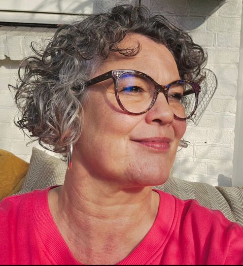 Foto van Els den Otter die opzij kijkt met krullend grijs haar, een bril en glimlacht voor een witte bakstenen muur.
