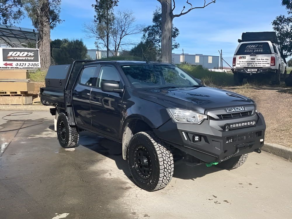 Dark gray Isuzu pickup truck with off-road modifications, parked outside with another truck in the background. β 100% Automotive In South Nowra, NSW