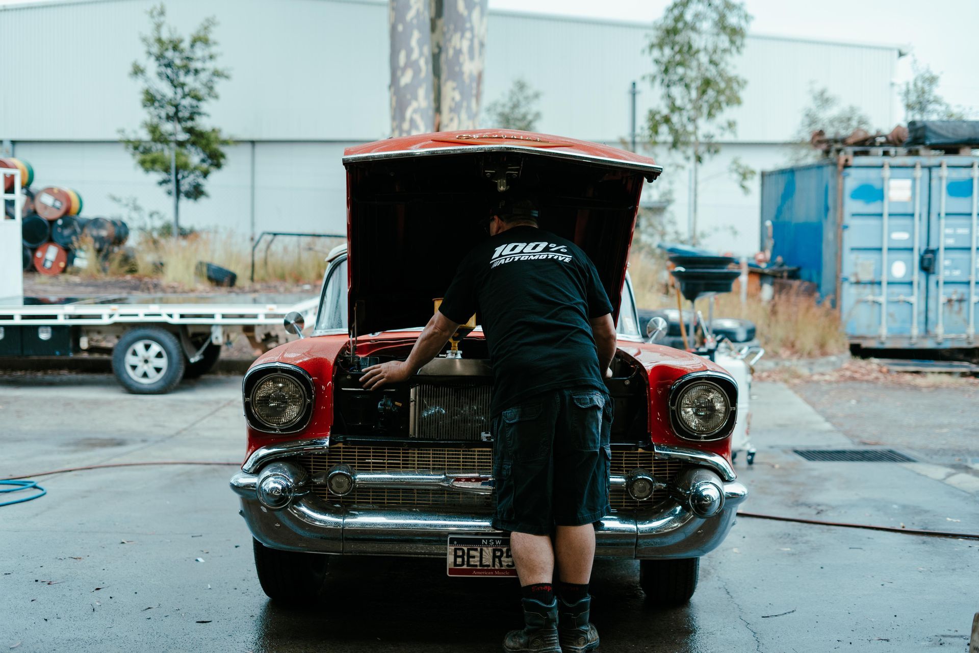 Person in black shirt working on a red classic car with open hood, in a wet outdoor setting. — 100% Automotive In South Nowra, NSW