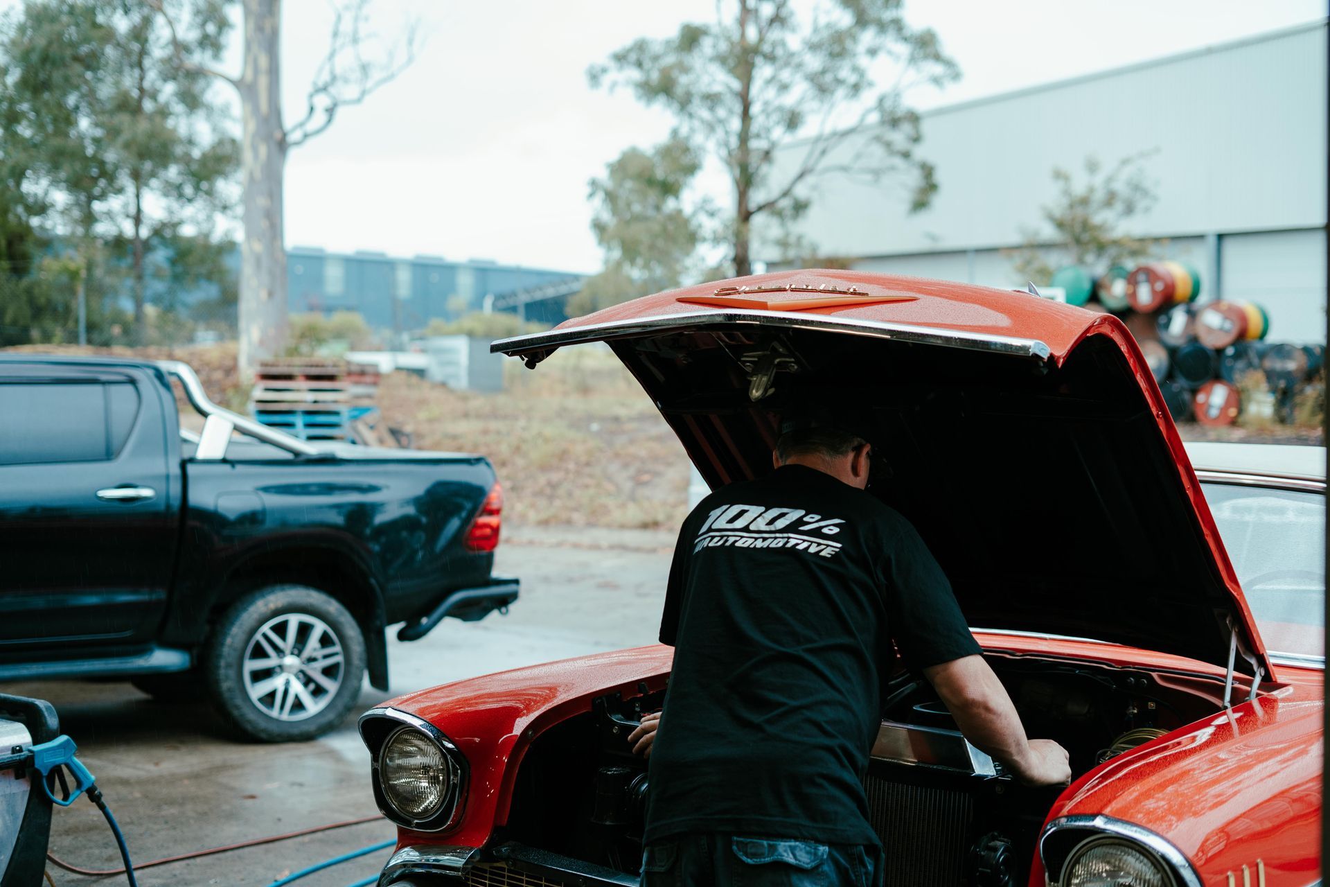 Man working on a red classic car with open hood, black truck parked beside. Outdoor setting. — 100% Automotive In South Nowra, NSW