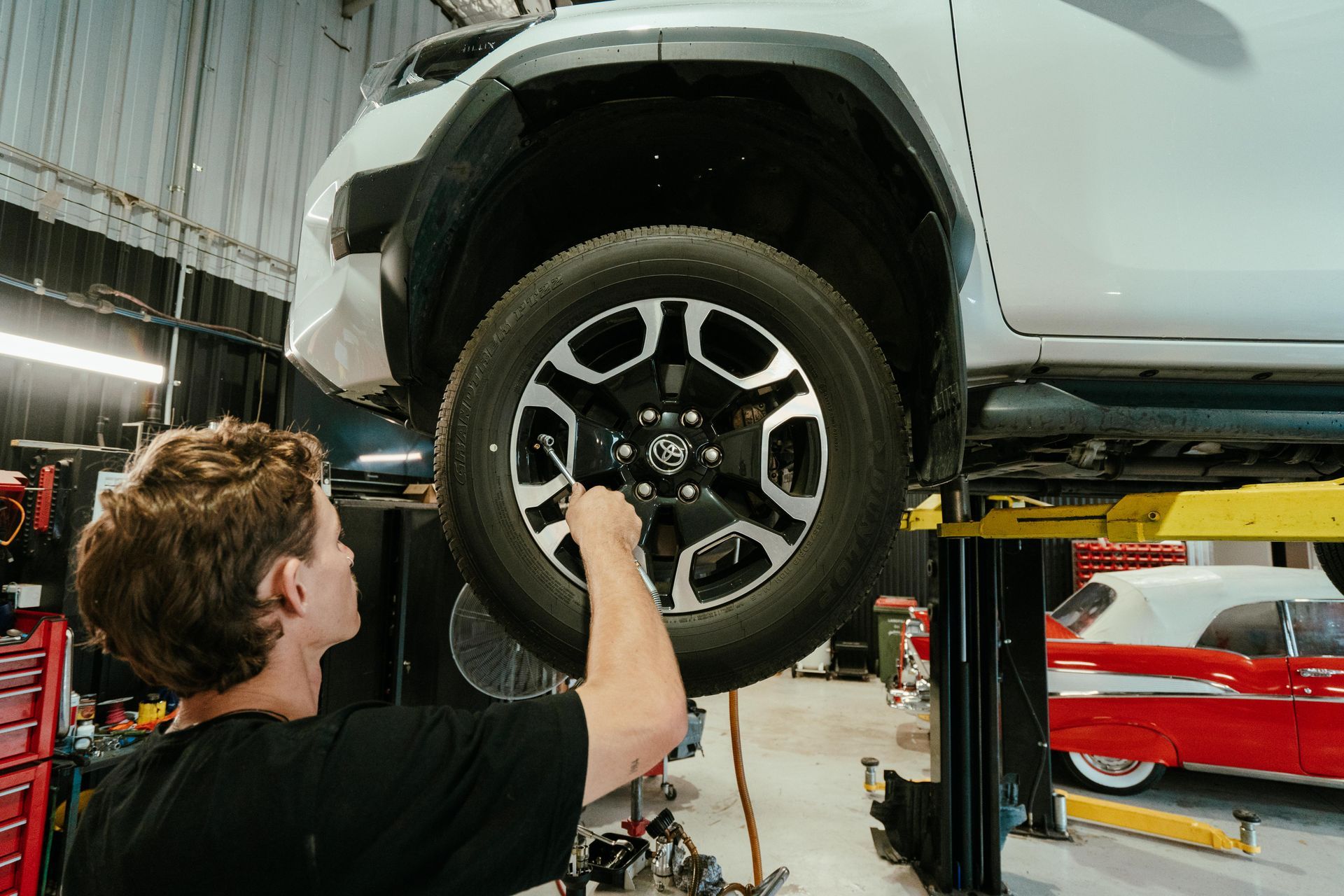 Mechanic working on the tire of a white truck in a garage; red toolbox and classic car visible. — 100% Automotive In South Nowra, NSW