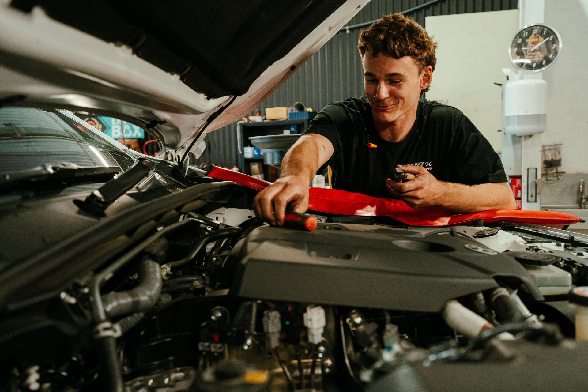 Mechanic working on car engine in a shop, smiling. Red tool, open hood. — 100% Automotive In South Nowra, NSW