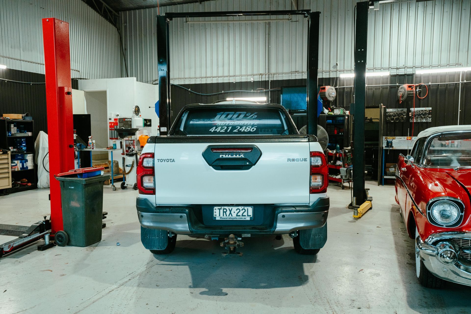 Rear view of a white Toyota Hilux truck in a garage, near a red classic car and a car lift. β 100% Automotive In South Nowra, NSW