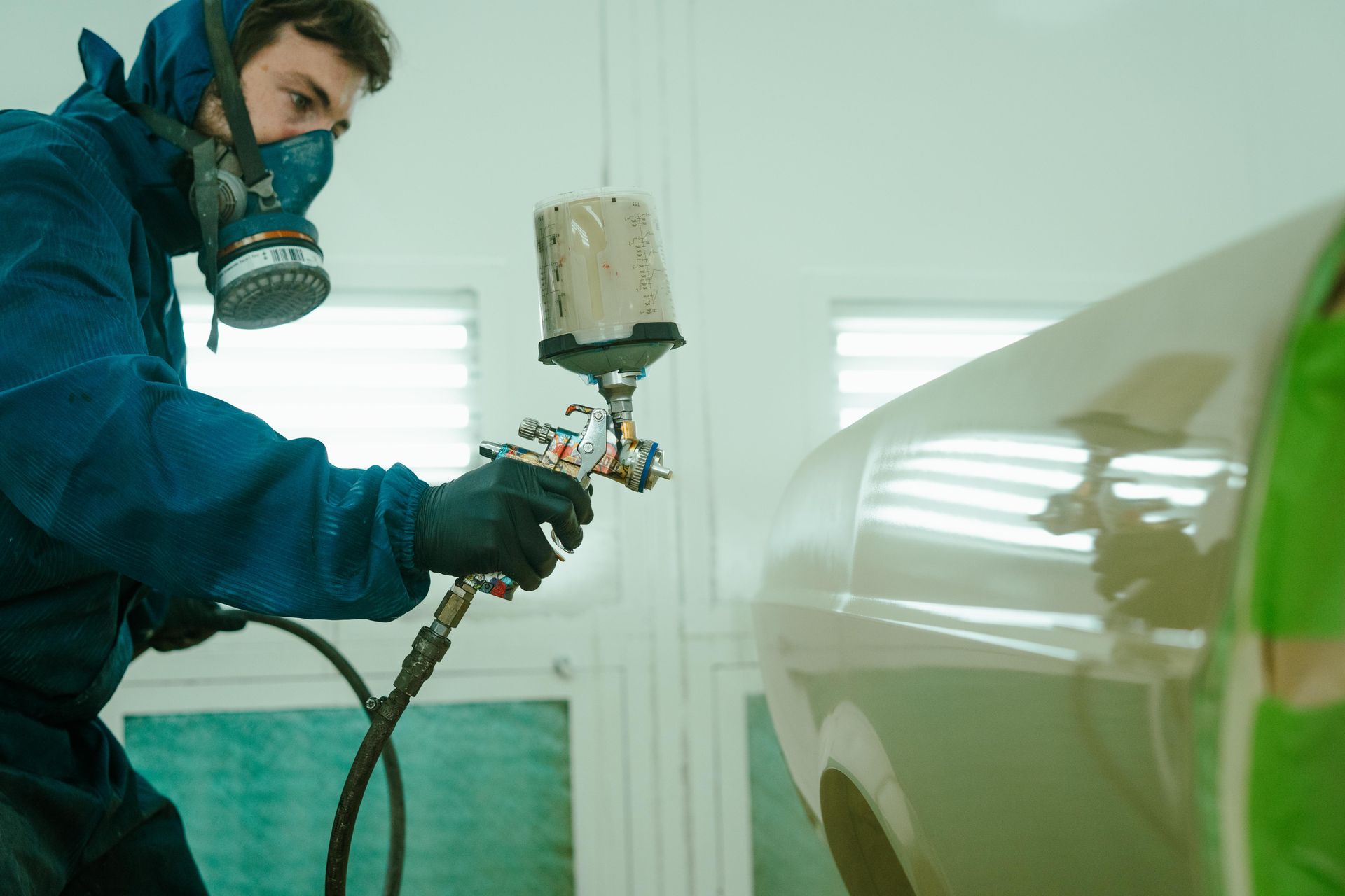 Person in blue protective suit and respirator spray painting a car in a light-colored workshop. — 100% Automotive In South Nowra, NSW