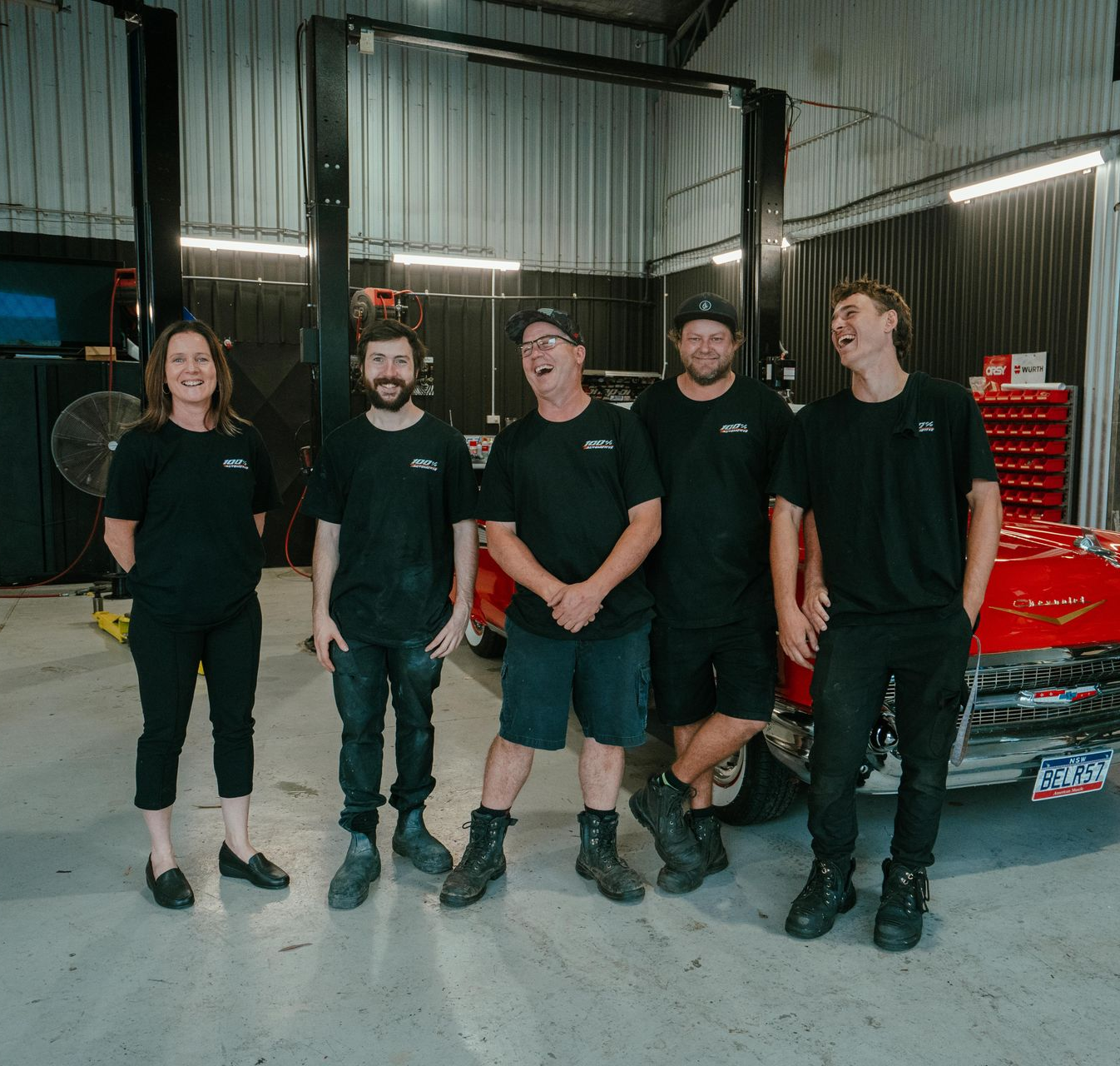 Five people in a car repair shop smiling; two men laughing near a red car, two others beside a vehicle lift. — 100% Automotive In South Nowra, NSW