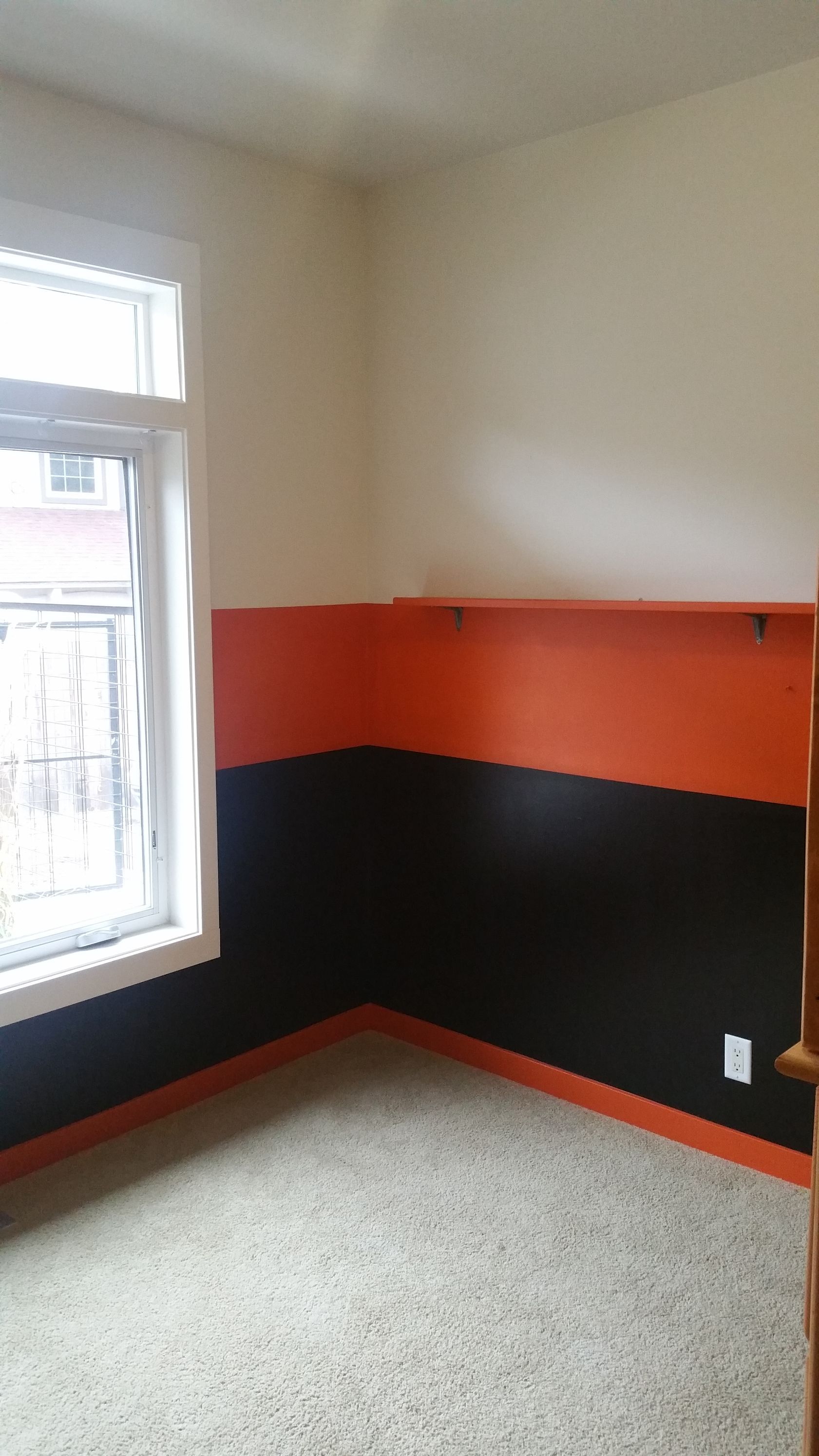 Empty corner of a room with a window, beige carpet, and black-and-orange painted walls.