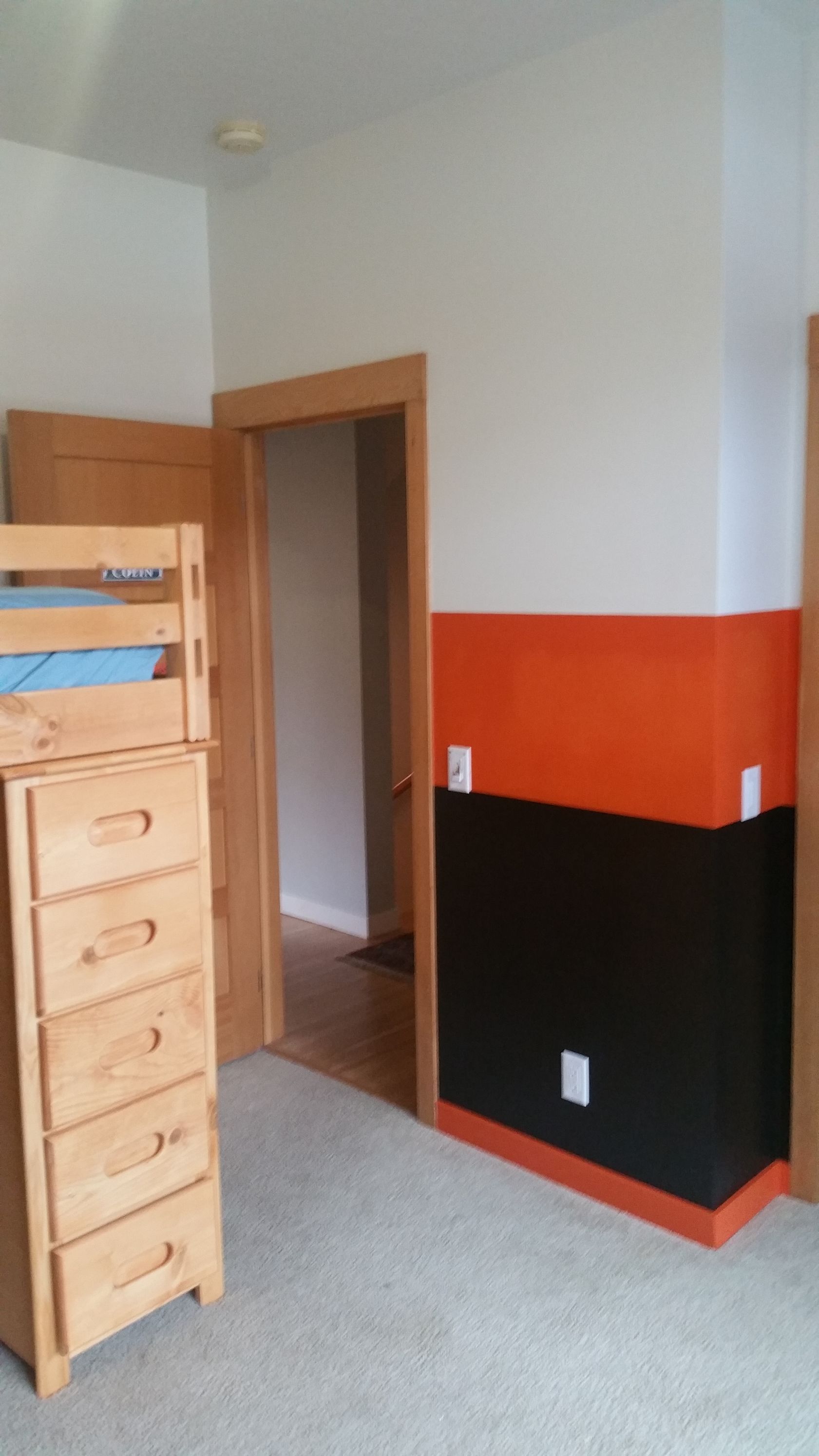 Small bedroom corner with a wooden dresser, open doorway, and black-and-orange painted walls.