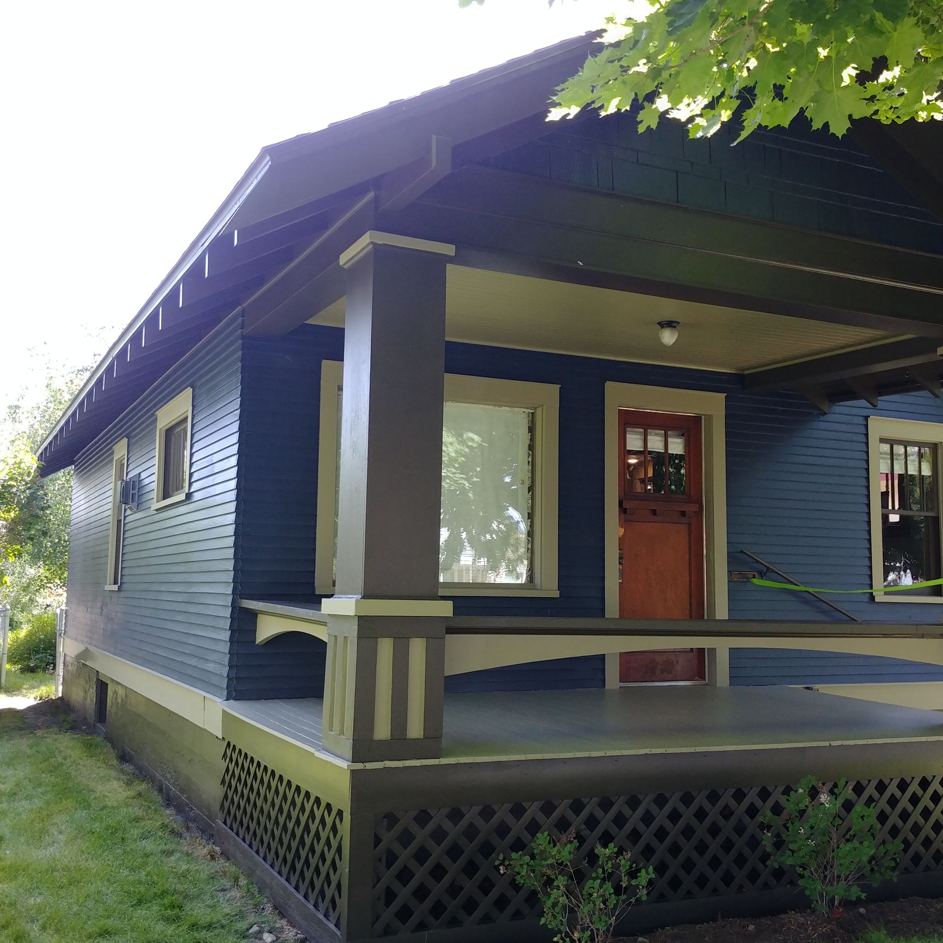 Blue house with a front porch, red door, and lattice skirting under trees