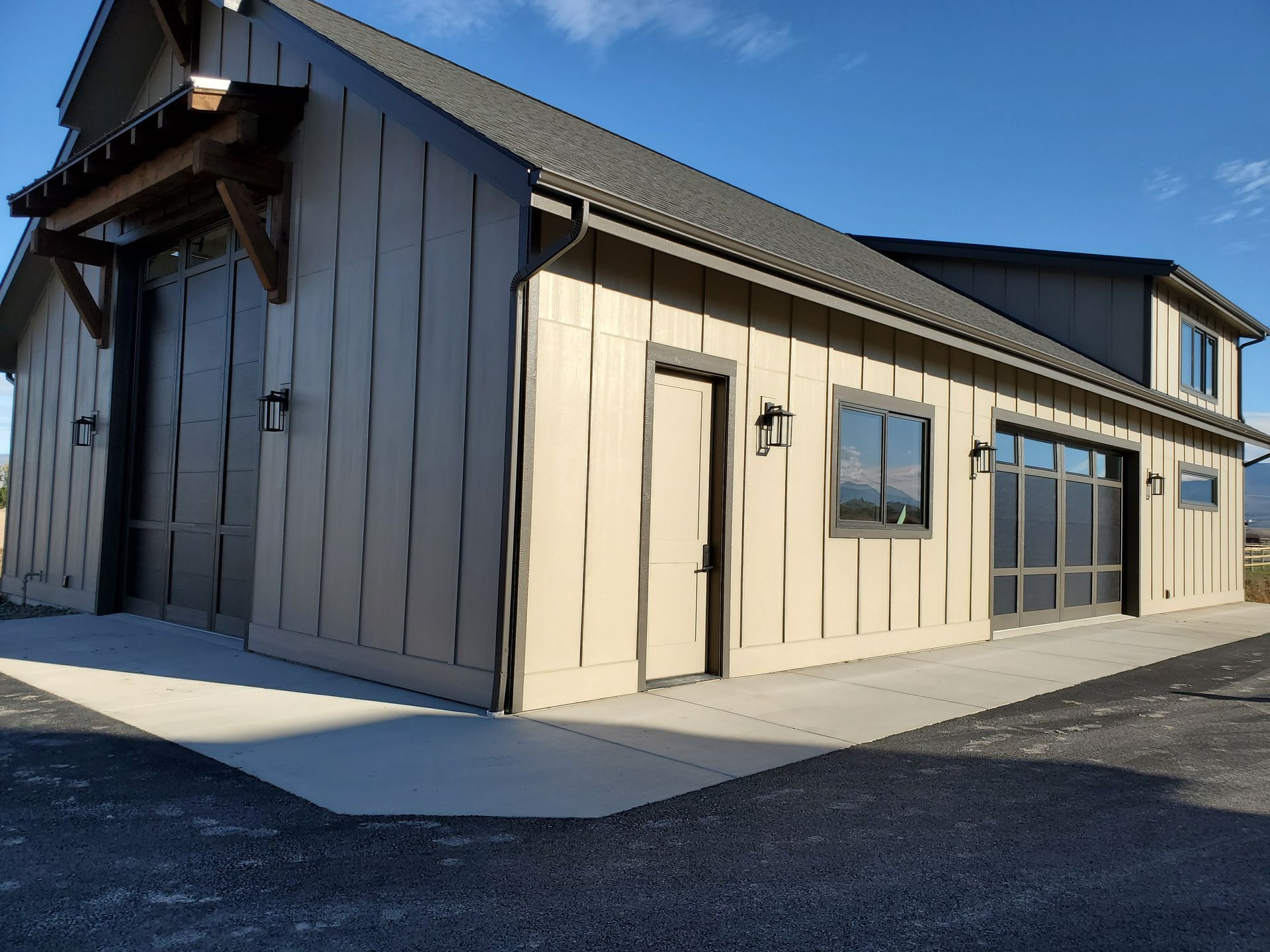 Modern beige barn-style building with dark roof and large garage doors on a gravel lot under a blue sky