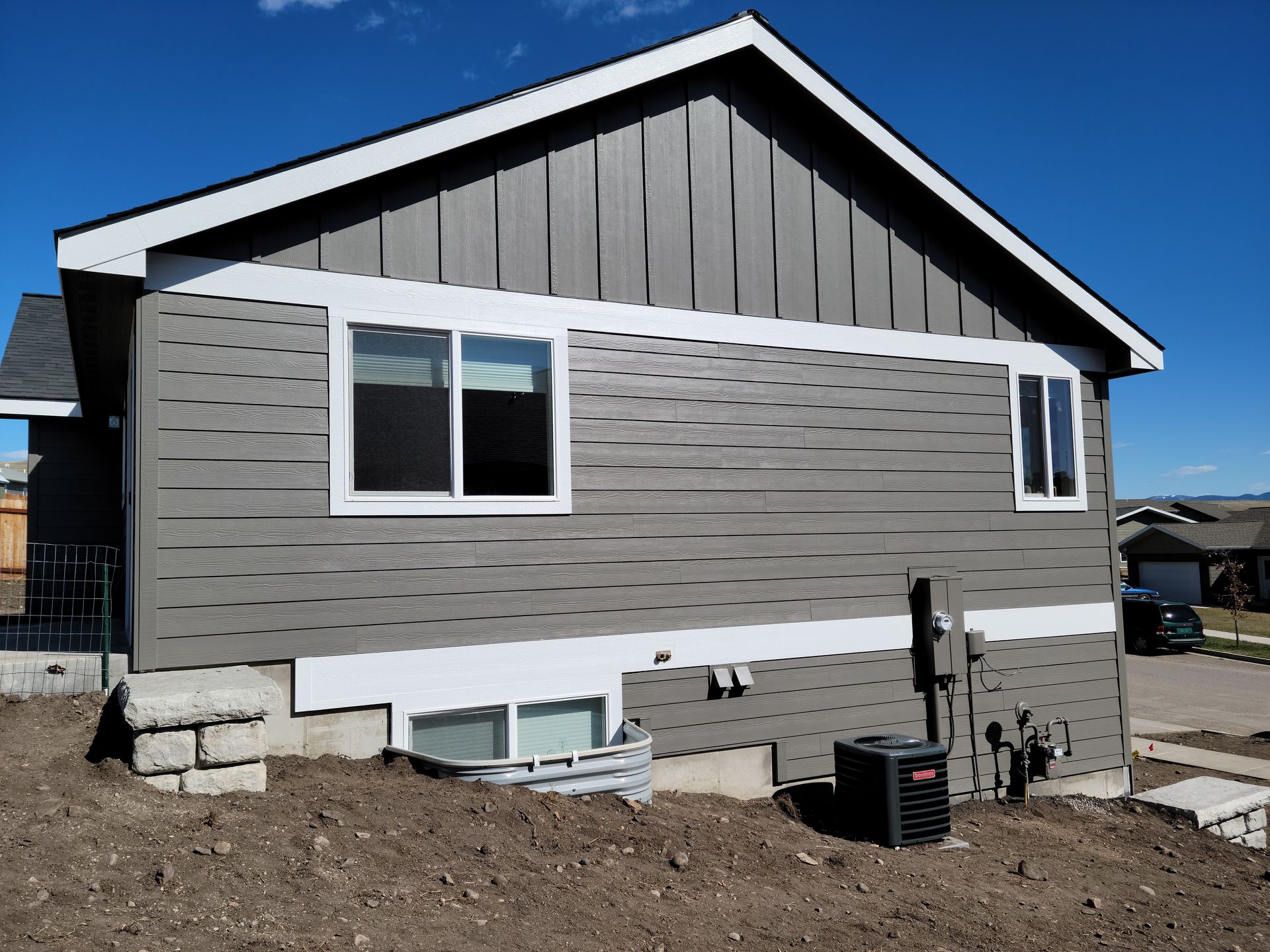 Gray house exterior with white-trimmed windows and dark gable siding on a bare dirt lot.