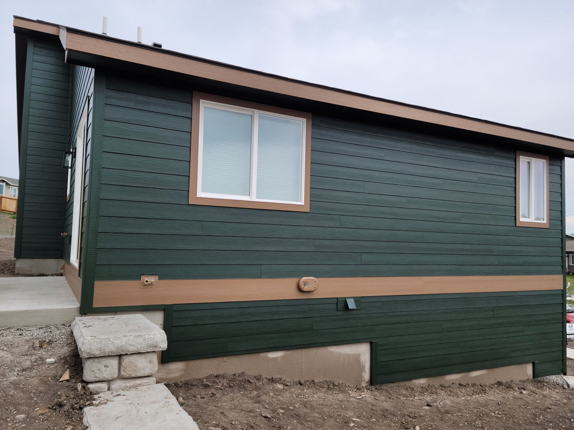Dark green house exterior with tan trim and a white-framed window, viewed from the side.