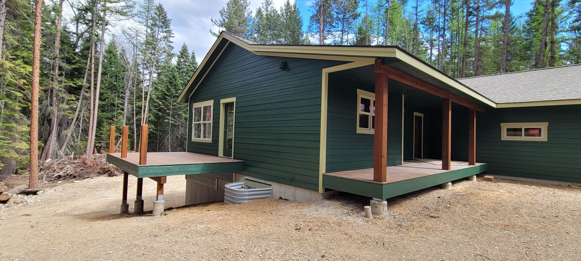 Green cabin in a pine forest with a small porch and gravel yard