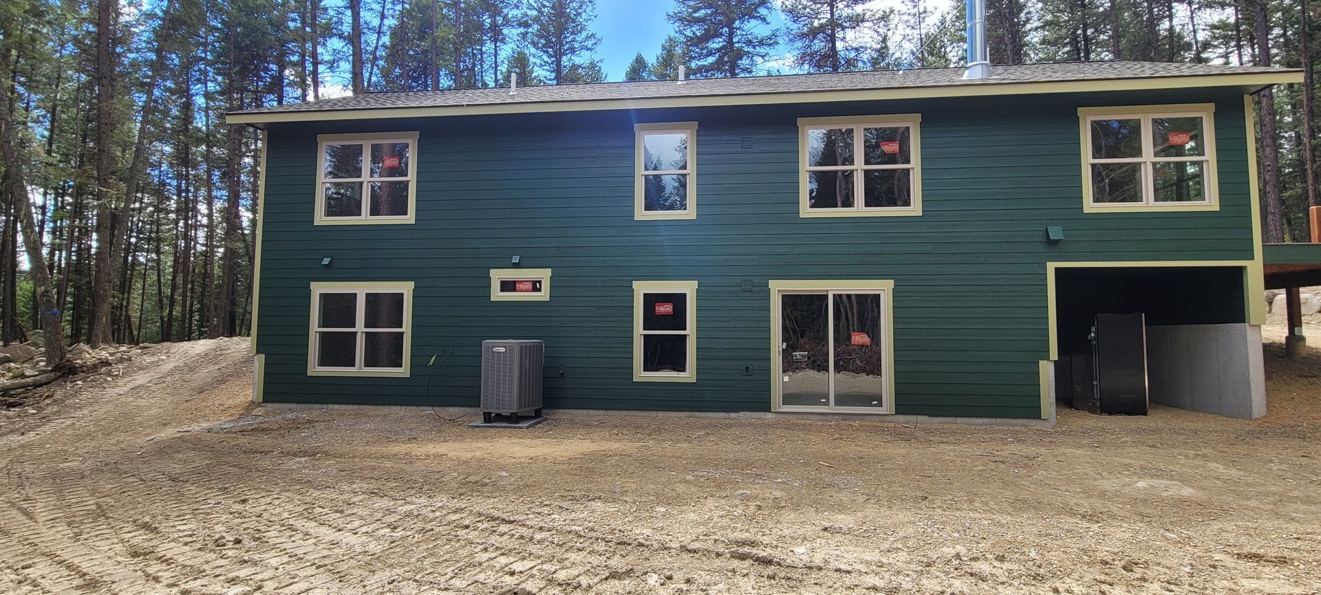 Green two-story house with white trim, set in a wooded area with a garage and dirt driveway.