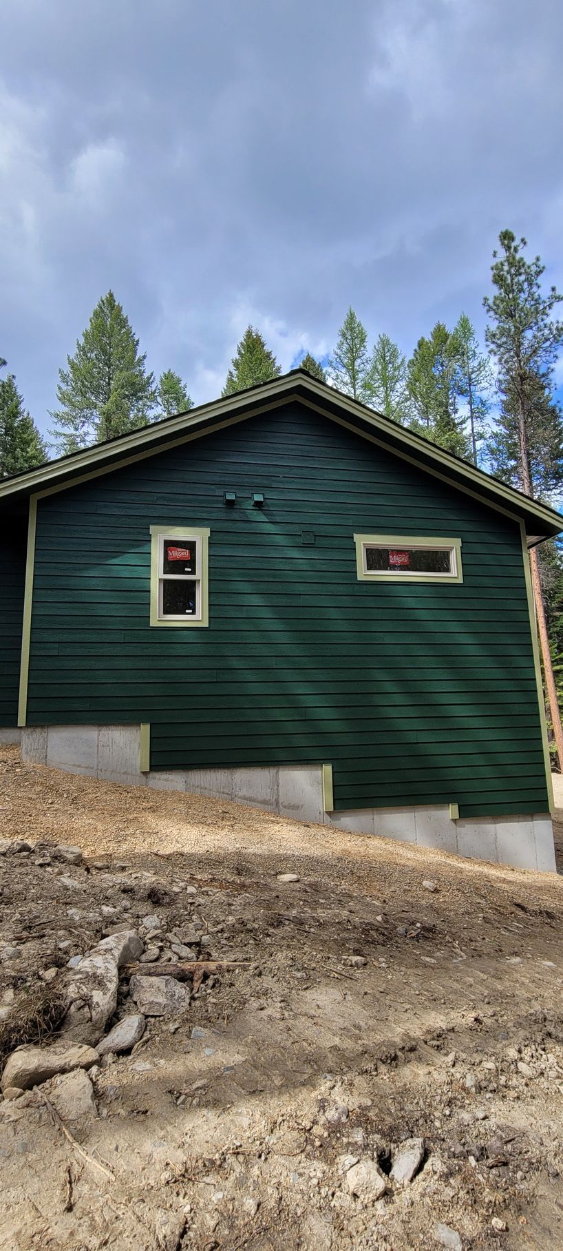 Green cabin on a rocky slope with pine trees and cloudy sky overhead