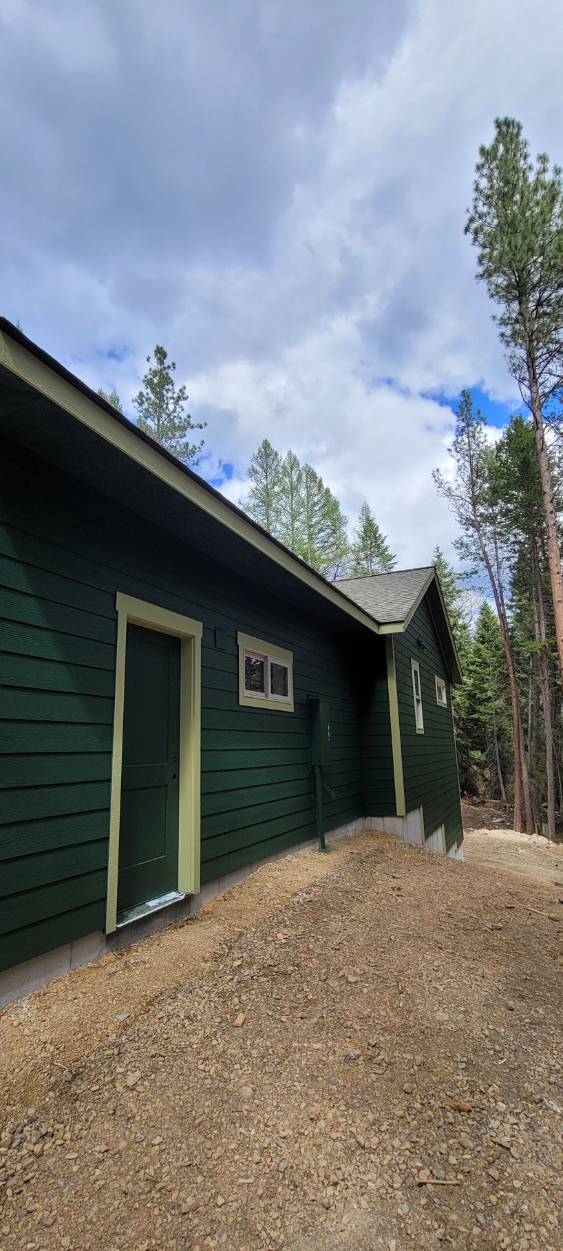 Dark green cabin exterior with a gravel path, tall pine trees, and a partly cloudy sky