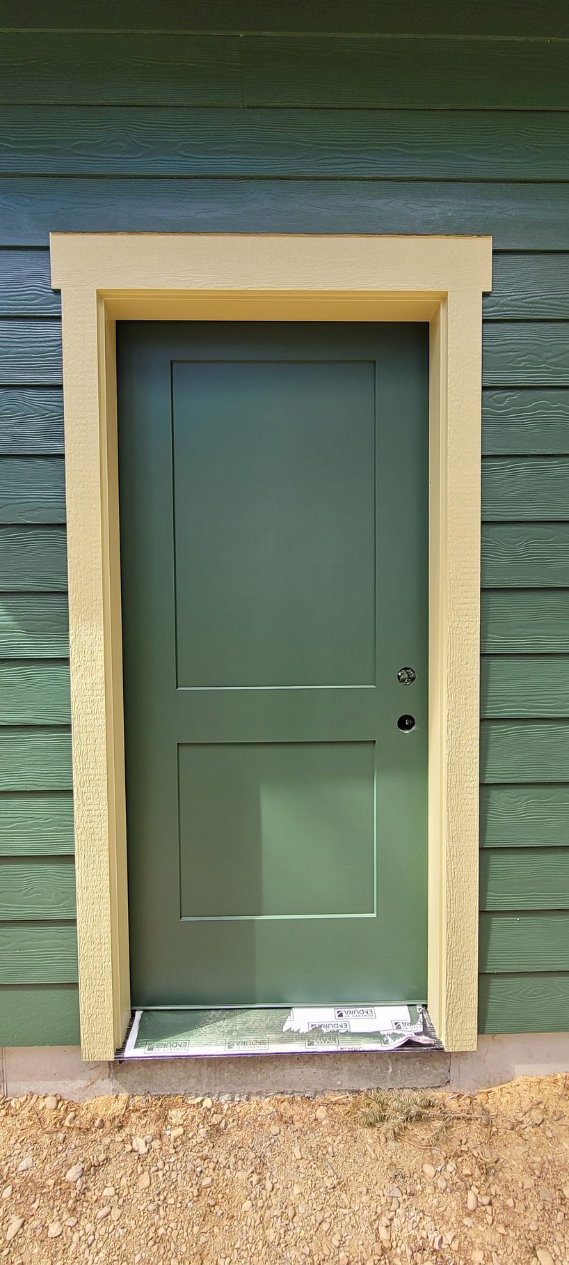 Green door with beige trim and a small damaged white threshold against a textured exterior wall