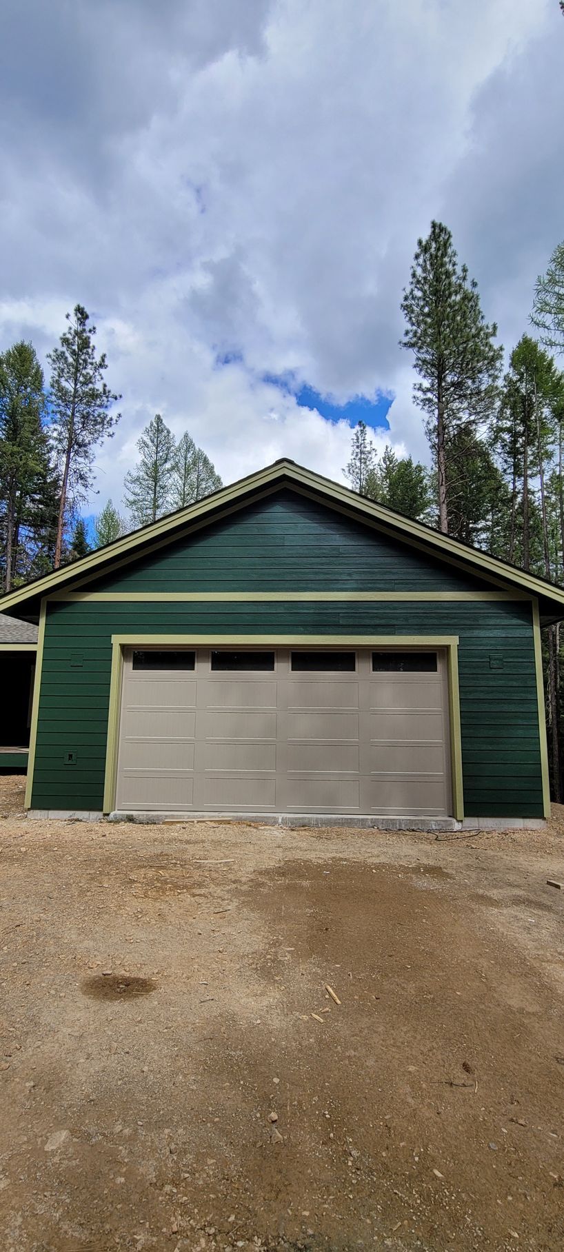 Green garage with beige door under a cloudy sky, framed by tall trees and a gravel driveway.