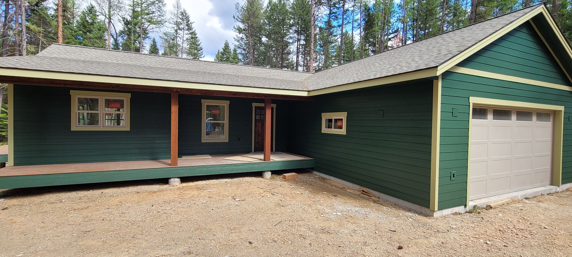 Green single-story house with brown trim, front porch, and attached garage in a wooded setting