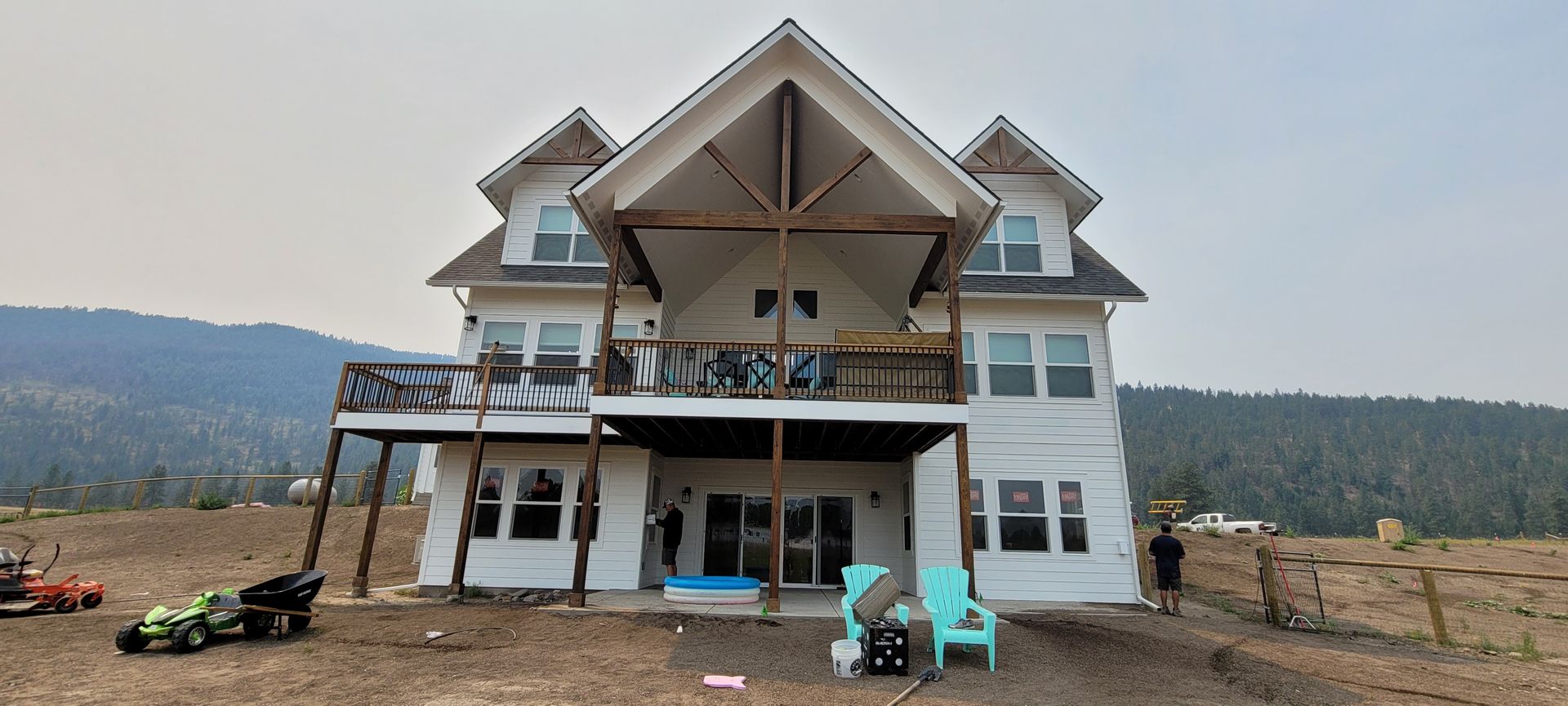 Large white house under construction with a front porch, in a rural field with scattered equipment.