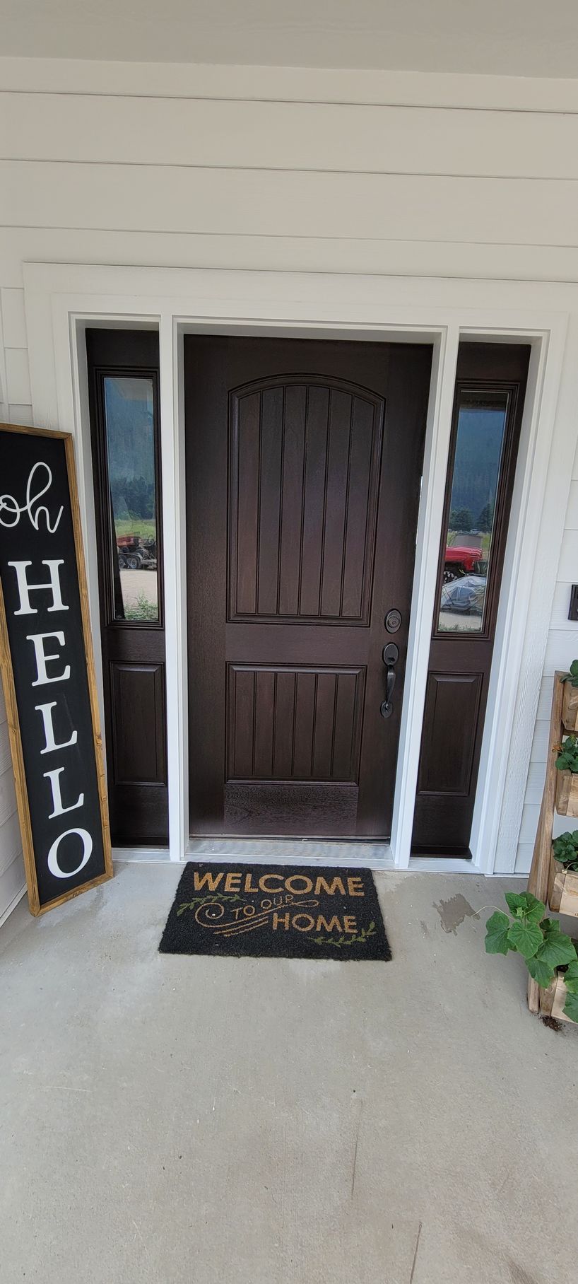 Front porch with a brown door, black HELLO sign, and welcome mat beside white siding.