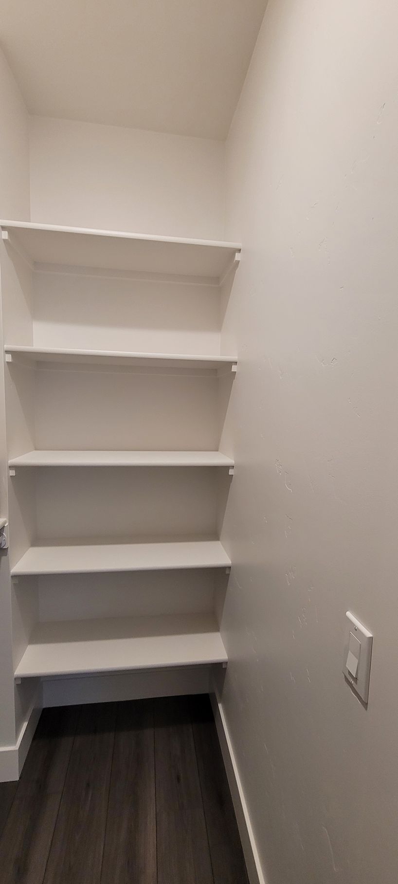 Small white shelving unit in a narrow closet with dark wood floor and light switch on the wall.