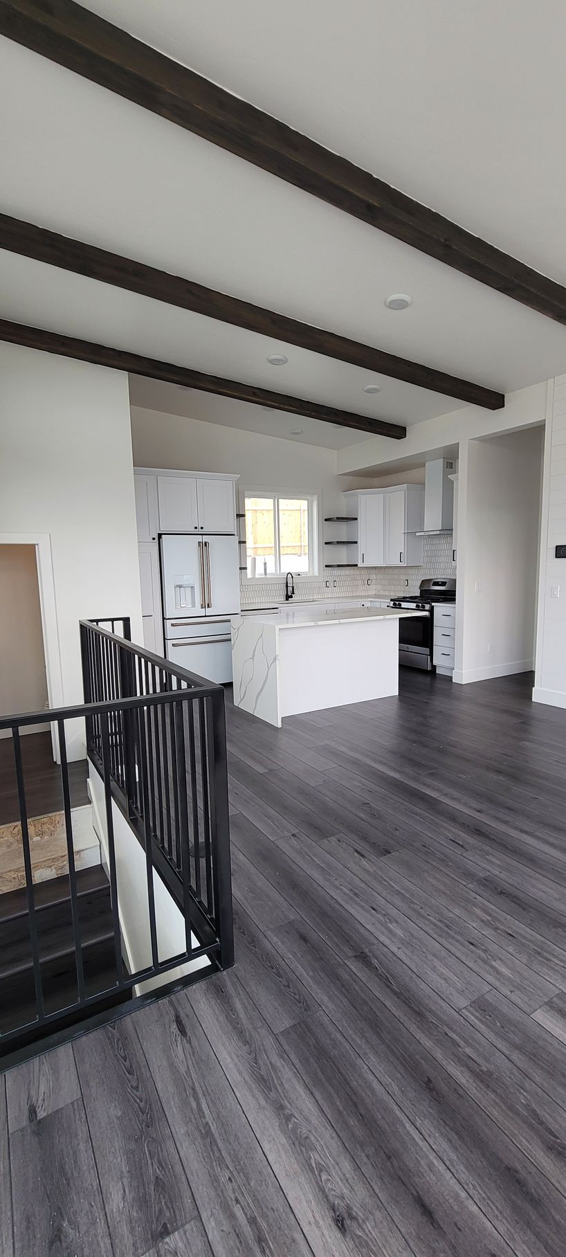 Modern open-plan interior with dark wood floors, white kitchen, exposed beams, and black stair railing