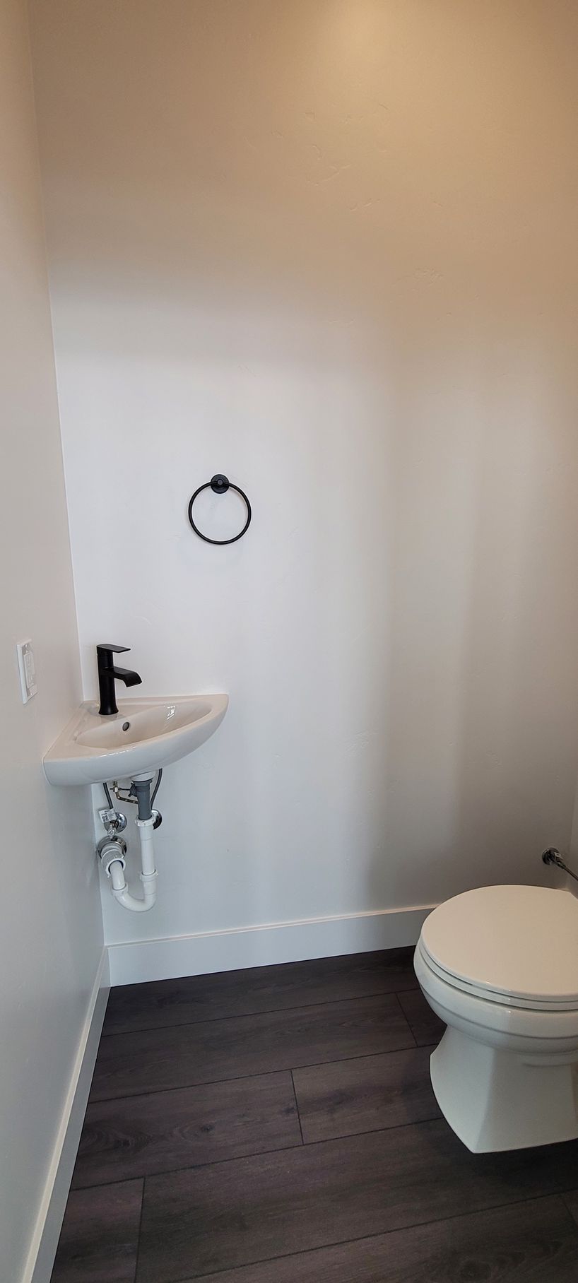 Small bathroom with white walls, pedestal sink, toilet, towel ring, and dark wood-look floor.