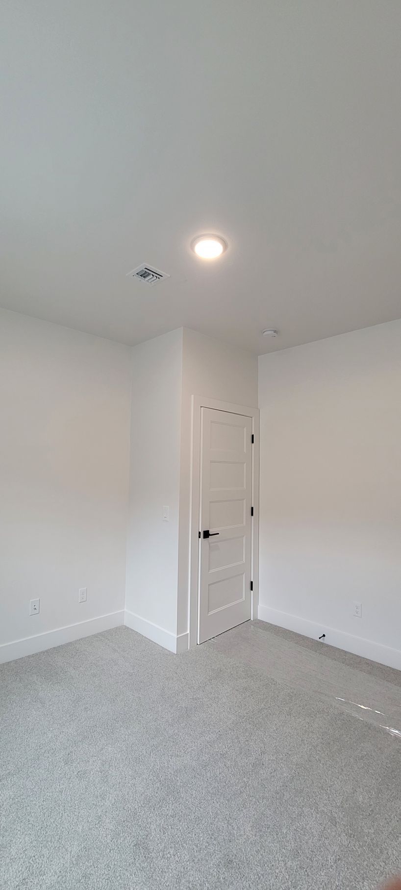 Small empty white bedroom with gray carpet, ceiling light, and a closed door.