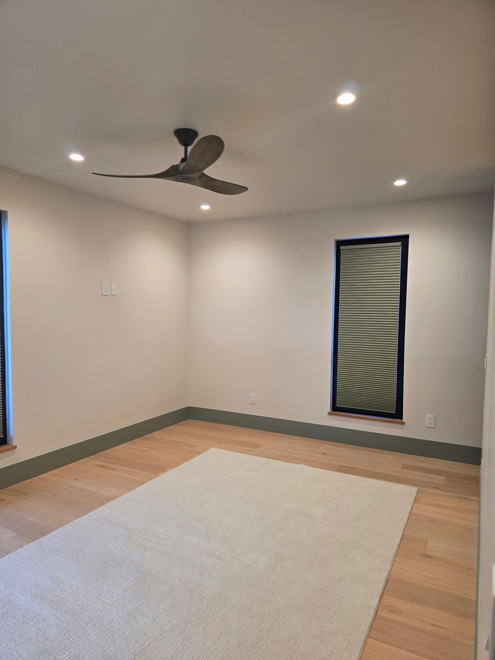 Empty modern room with ceiling fan, recessed lights, wood floor, large rug, and narrow windows with blinds