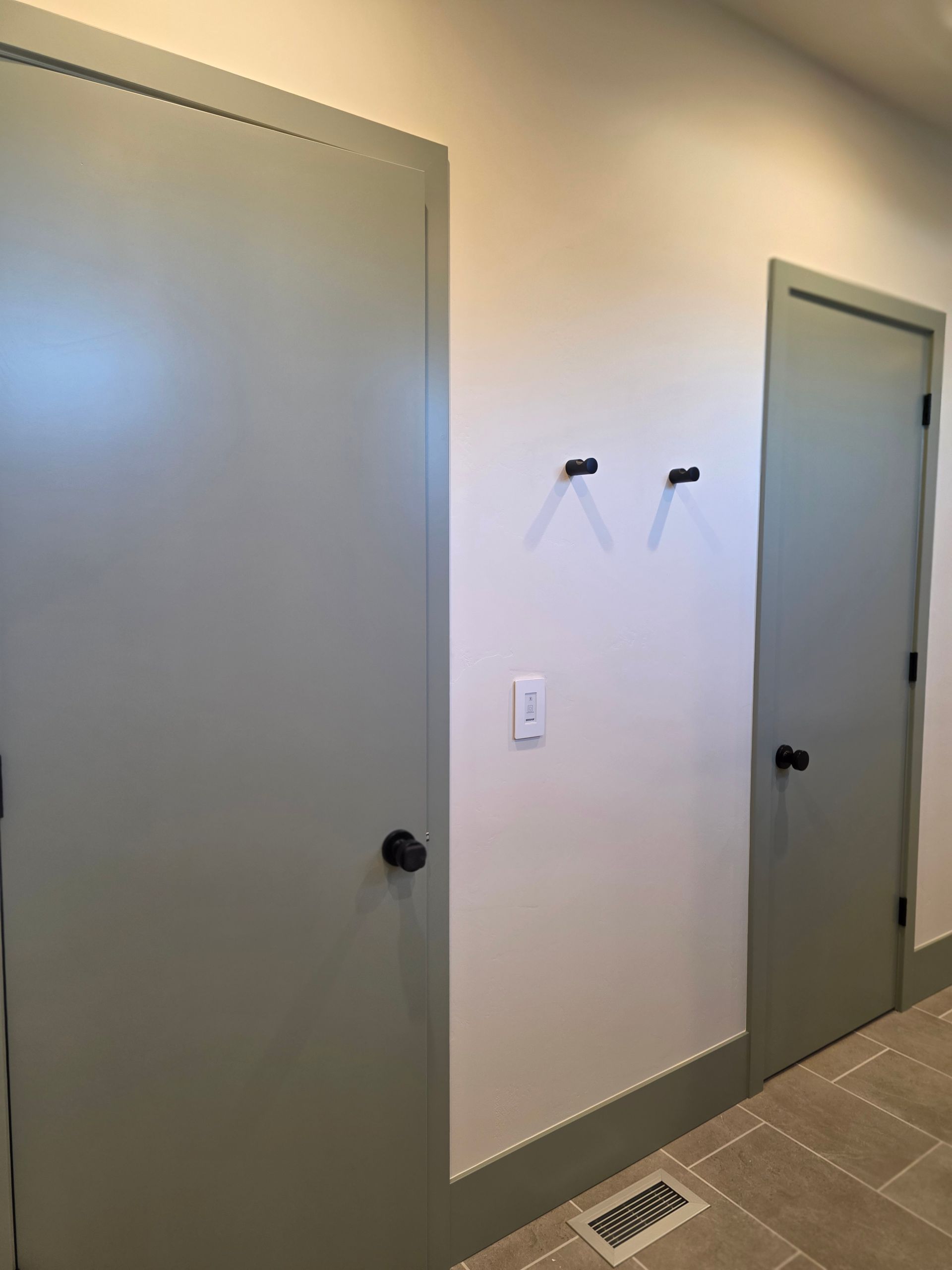 Hallway with two gray doors, two wall hooks, and a floor vent on tiled floor