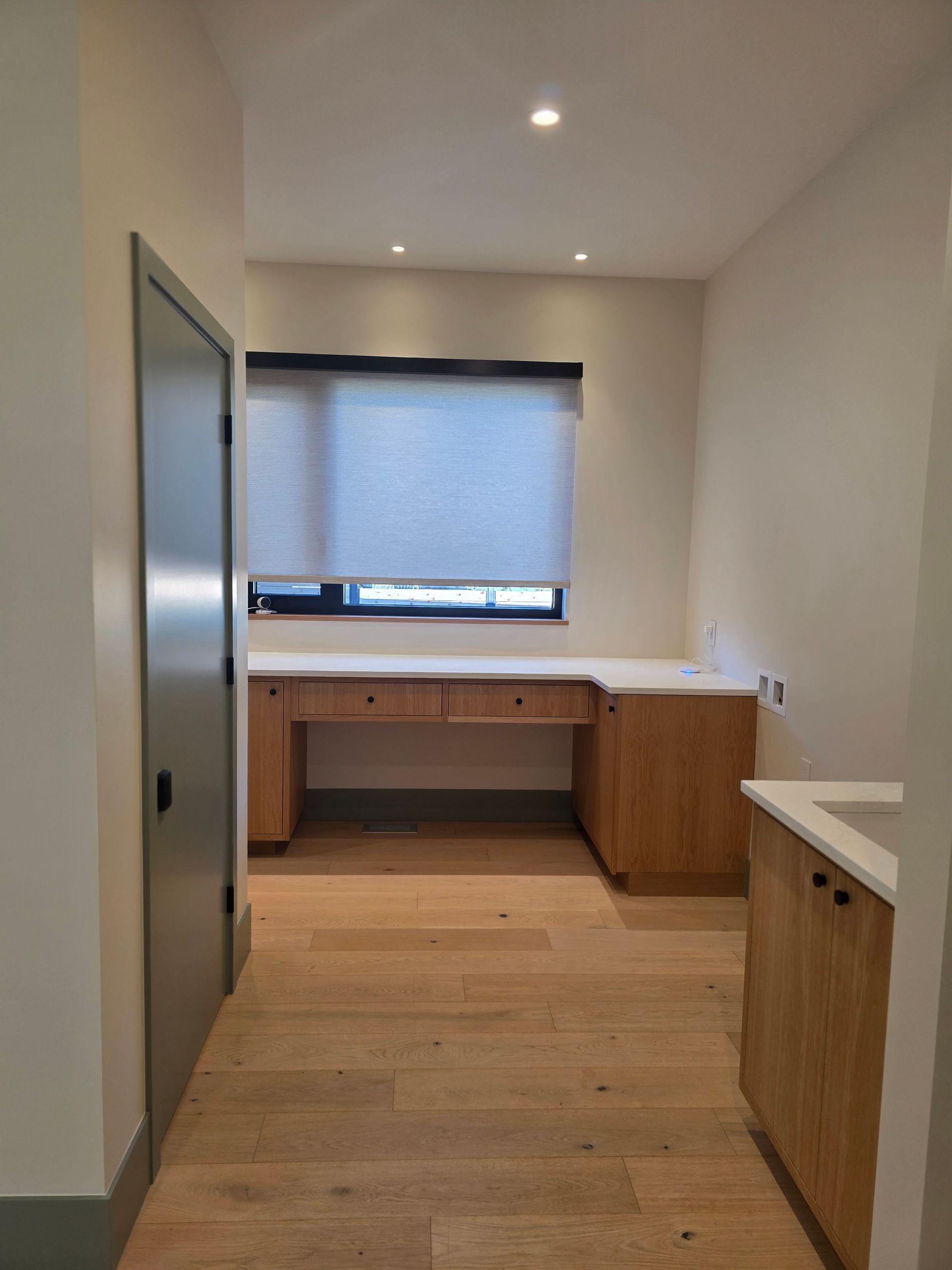 Narrow modern kitchen with light wood cabinets, white walls, and a window with a closed blind.